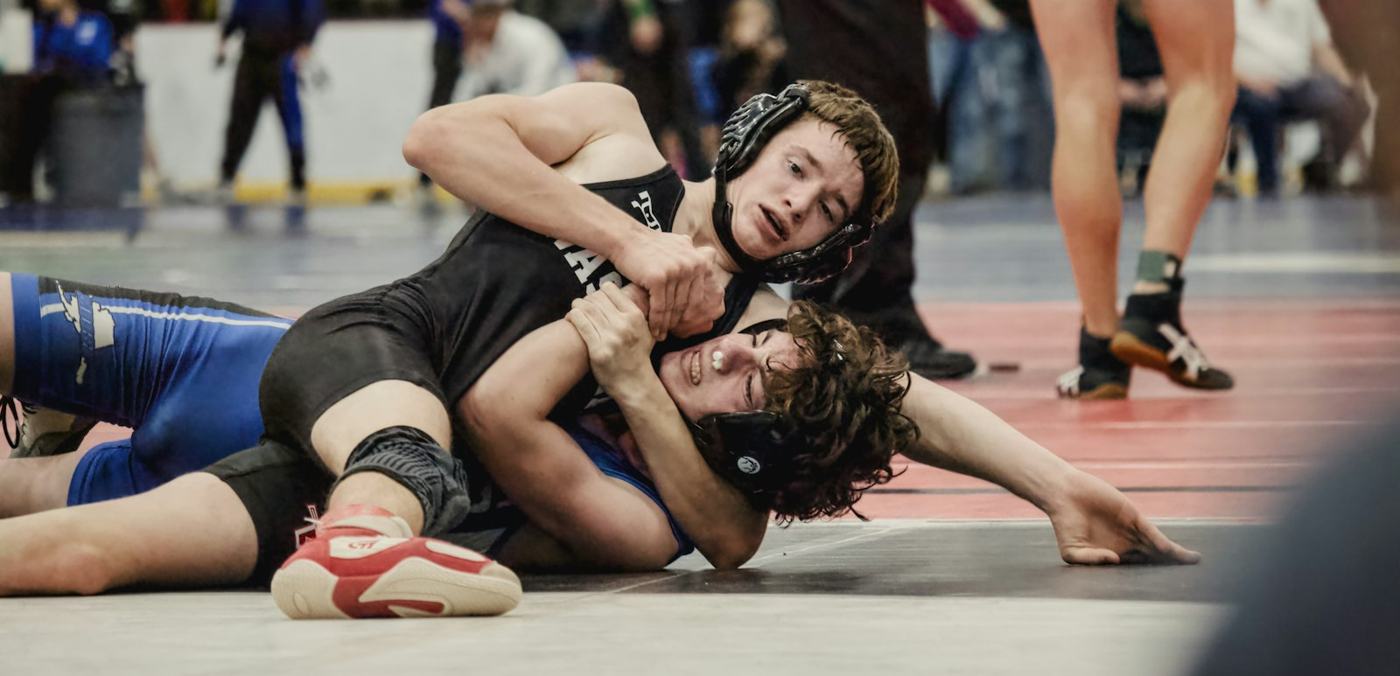 Two wrestlers on the mat with a coach watching